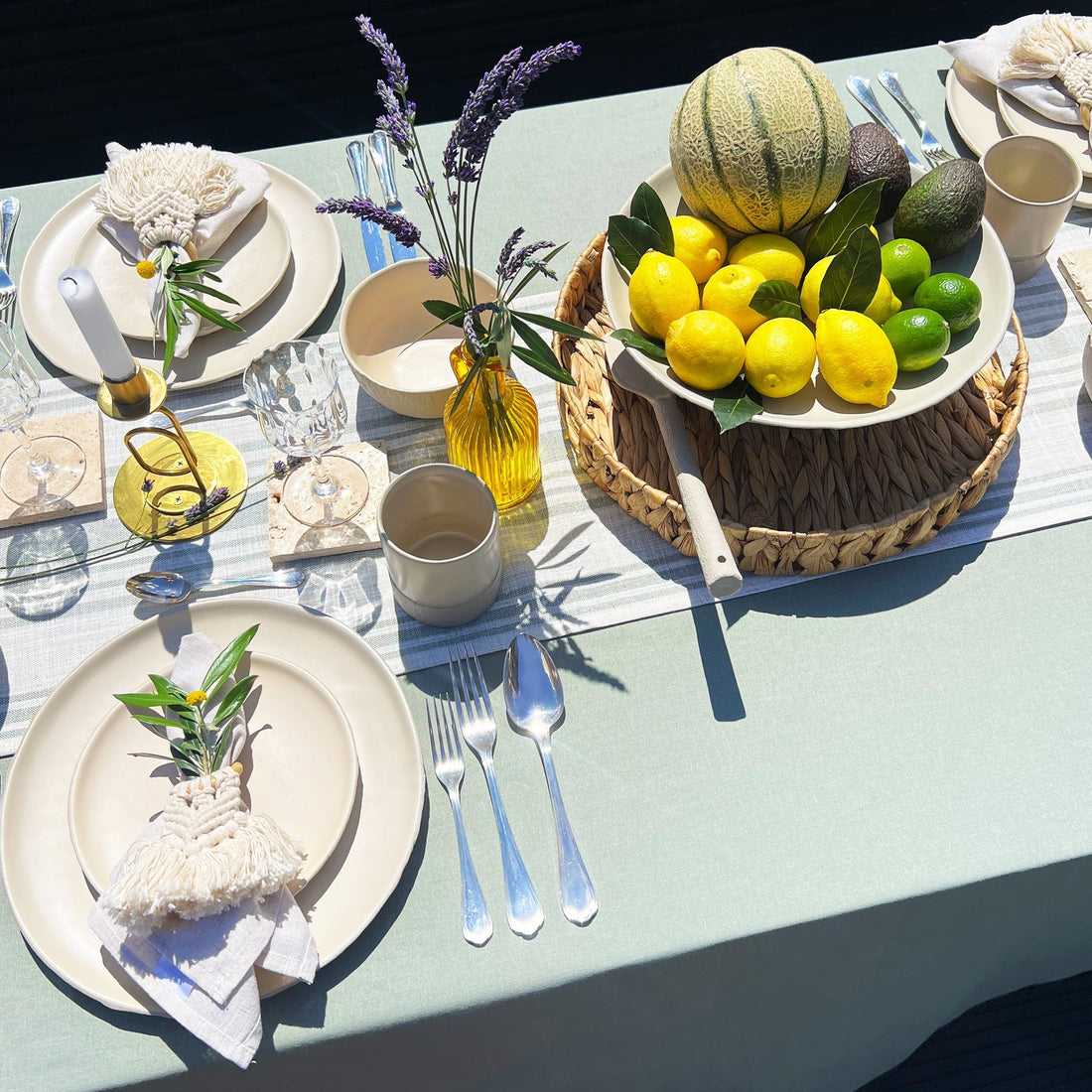 Beautiful al fresco dining table featuring beige handmade ceramic dinnerware, sage linen napkins, olive branches, lavender, and a citrus fruit centerpiece on a ceramic tray.