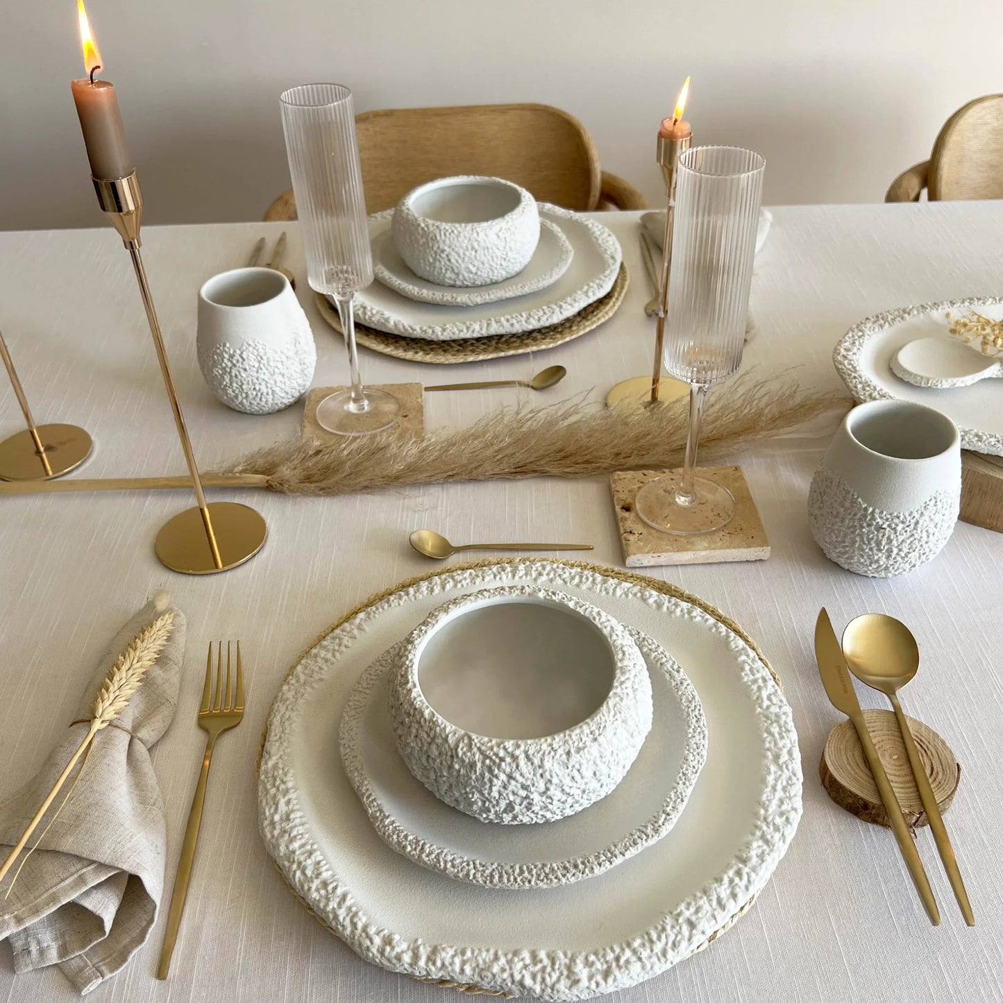 A styled dining table setting with textured white ceramic bowls, plates, bowls with matching serving platter, paired with gold-toned flatware, candlesticks, and neutral linens in a warm, rustic-chic interior.