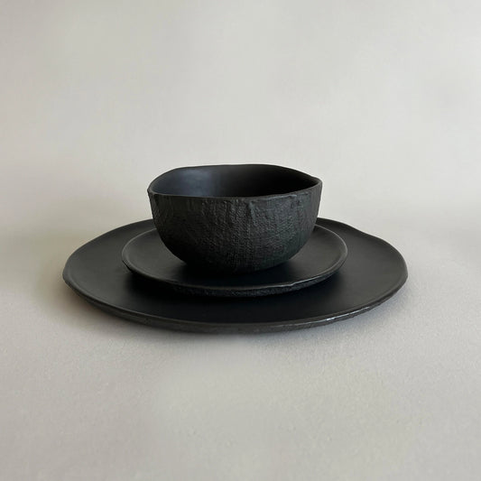 A black textured ceramic bowl set with large dinner plate and side plate, showcasing organic linen texture.