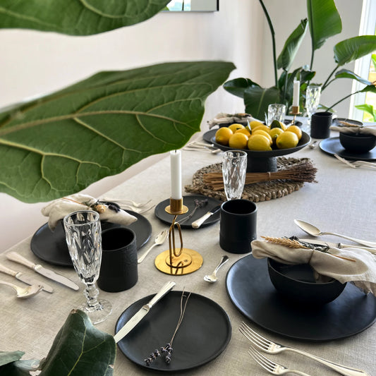Elegant table setting with black matte ceramic dinner plates, dessert plates, soup bowls, cups, silverware, and glasses on a beige linen tablecloth.