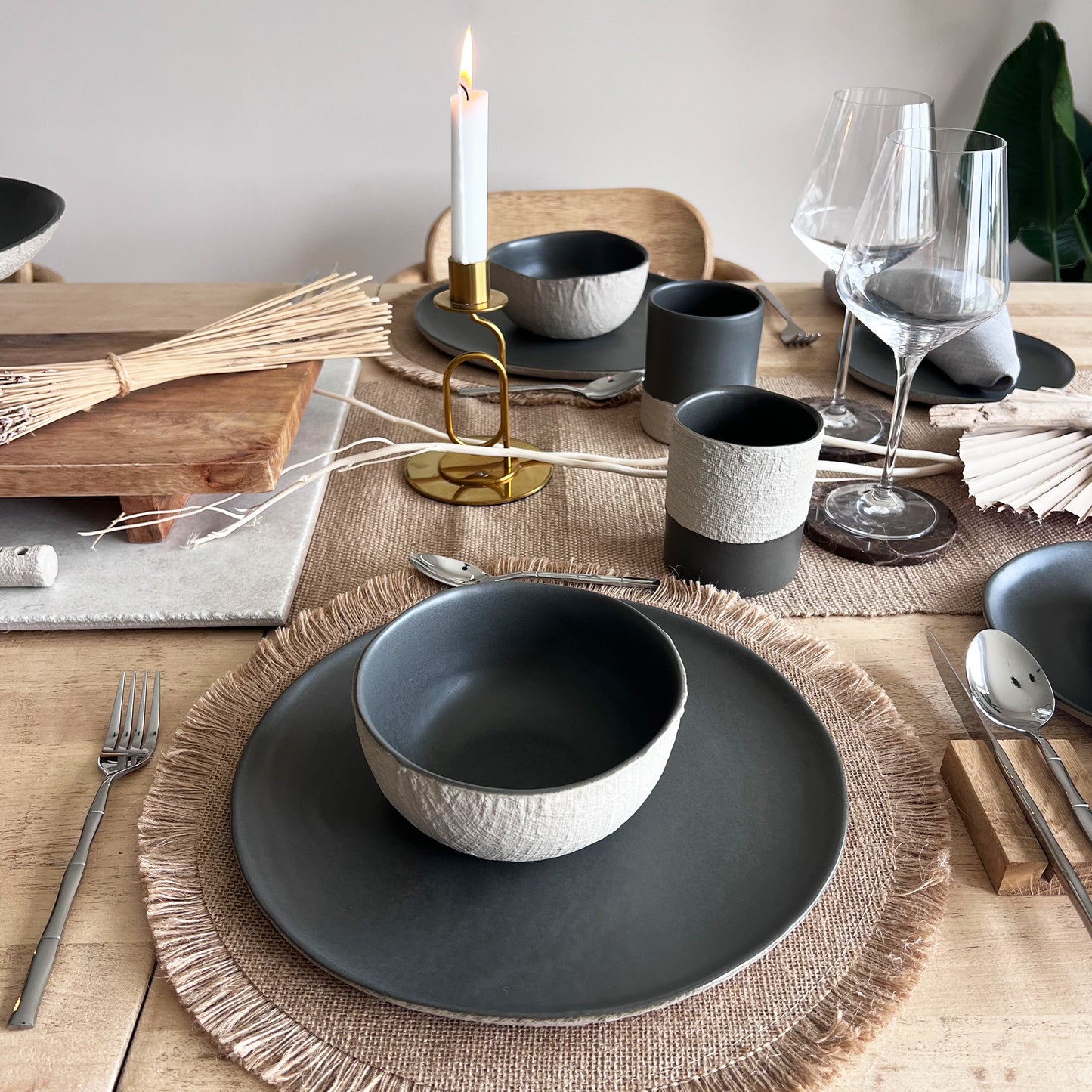 Dining table setting with beige and black ceramic bowls, glasses, plates, mugs and cutlery on a wooden table.