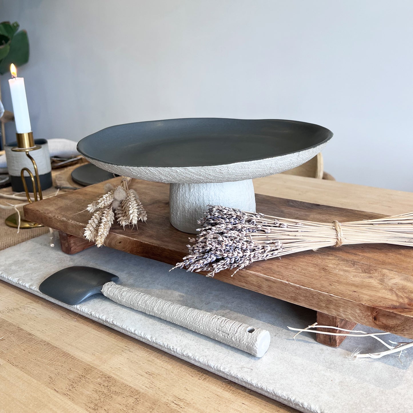 Decorative setting with a dark grey ceramic centrepiece on a wooden stand, with ceramic serving spoon, and dried plants.