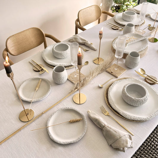 Elegant table setting with white ceramic plates, gold cutlery, and candles on a white linen tablecloth.