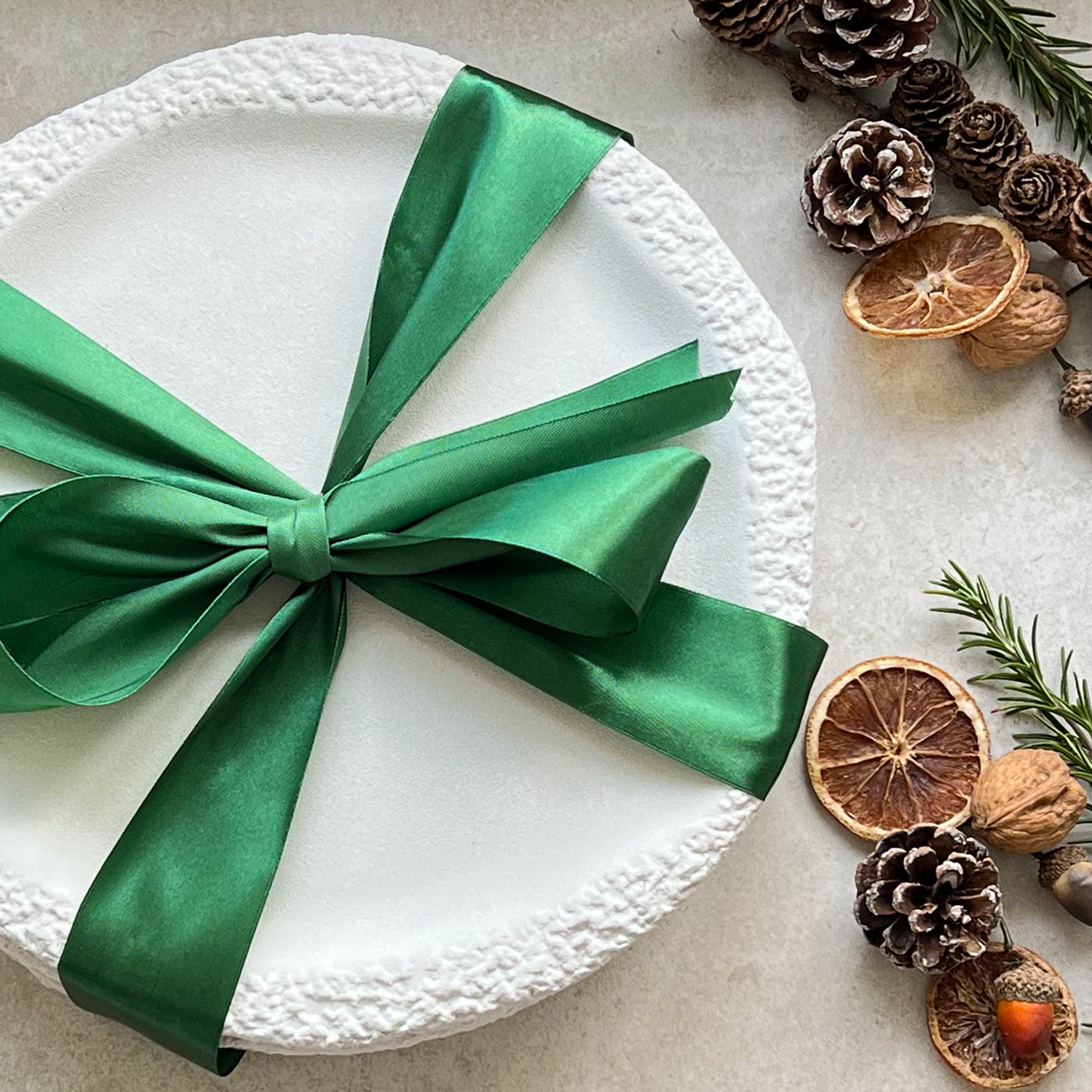 White ceramic dinnerware set plate with a green ribbon, surrounded by pine cones and dried oranges .