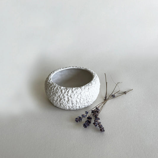 A small round textured white ceramic cereal bowl with a rough, pebbled exterior, shown on a light grey surface with a small sprig of dried lavender beside it.