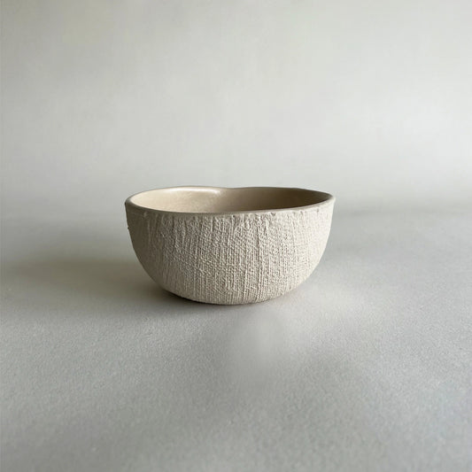 A organic-shaped ceramic bowl with a textured, ribbed exterior and a smooth, matte beige interior, set against a neutral light grey background.