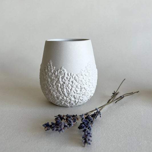 A matte white ceramic coffee cup with a textured, rougher pattern around the lower half, styled with dried lavender sprigs on a neutral light beige background.