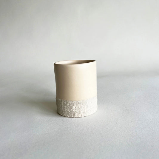 A minimalist handmade ceramic cup with a smooth, matte cream-coloured upper body and a textured, lighter beige lower band, set against a neutral light grey background.
