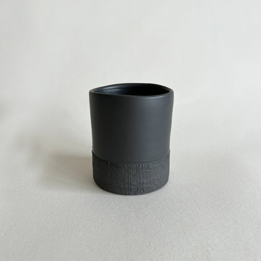 A minimalist matte black ceramic cup with a textured, woven band around the lower half, set against a clean, light grey background.