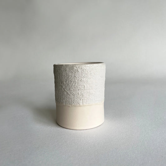 A cylindrical ceramic textured coffee mug, with textured organic upper half and a smooth, matte cream lower half, set against a neutral grey background.