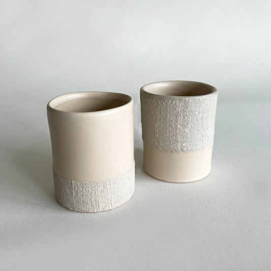Two large ceramic cups in a warm cream colour, featuring a smooth matte finish on the upper body and a textured, ribbed band near the base, set against a neutral light grey background.