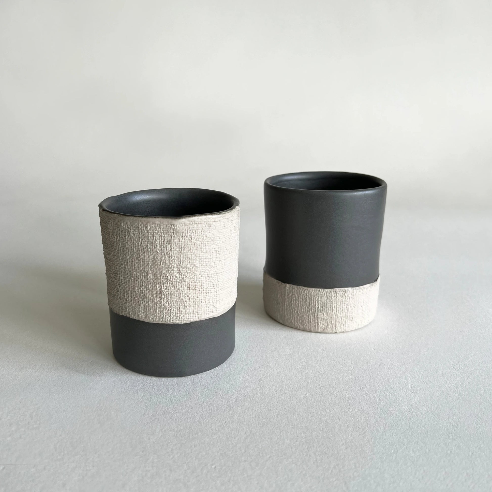 A set of two coffee cups and mugs organically shaped, in matte charcoal colour with beige contrasted textured woven finish. 