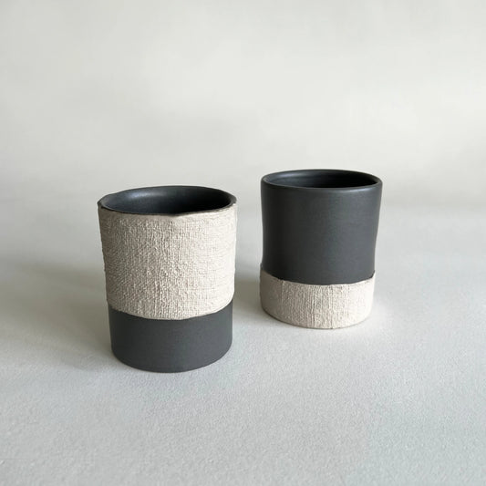 A set of two coffee cups and mugs organically shaped, in matte charcoal colour with beige contrasted textured woven finish. 