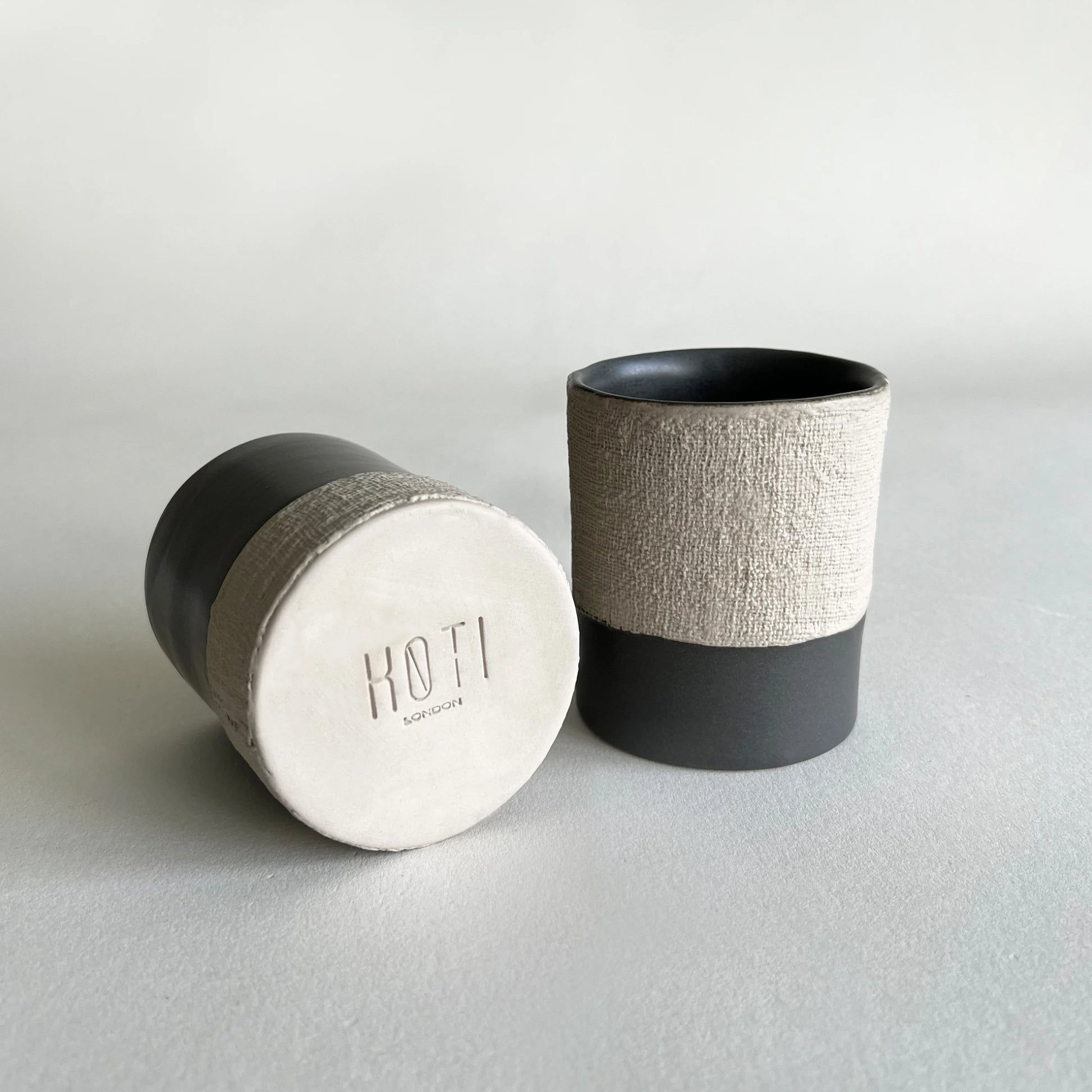 Two cups and mugs in ceramic material, handmade, in matte charcoal colour contrasted with beige organic texture.