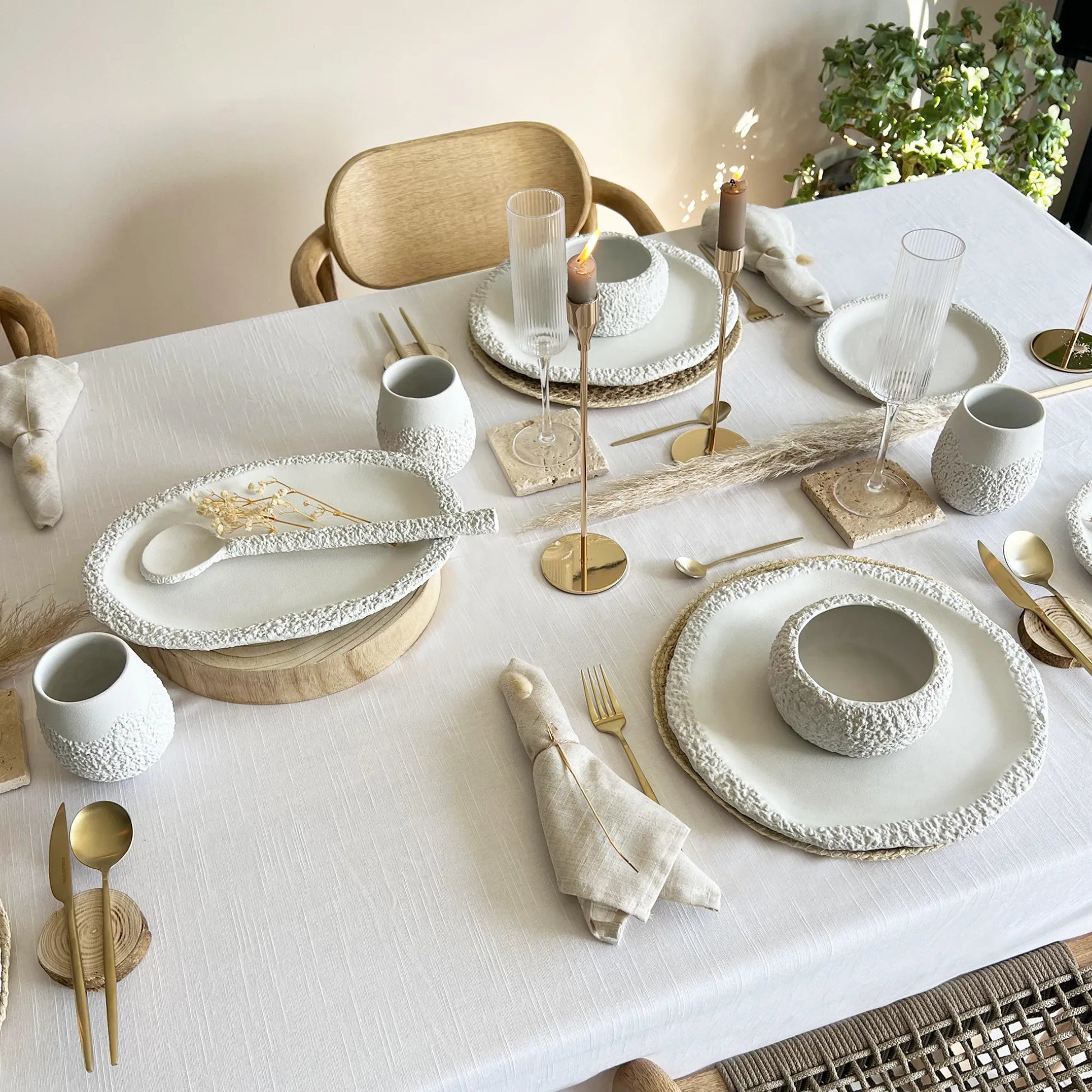 A styled dining table setting with textured white ceramic plates, matching bowls, cups, serving tray and spoons, gold toned cutlery and candlesticks, linen napkins, and neutral table runners on a white tablecloth in a bright indoor room.