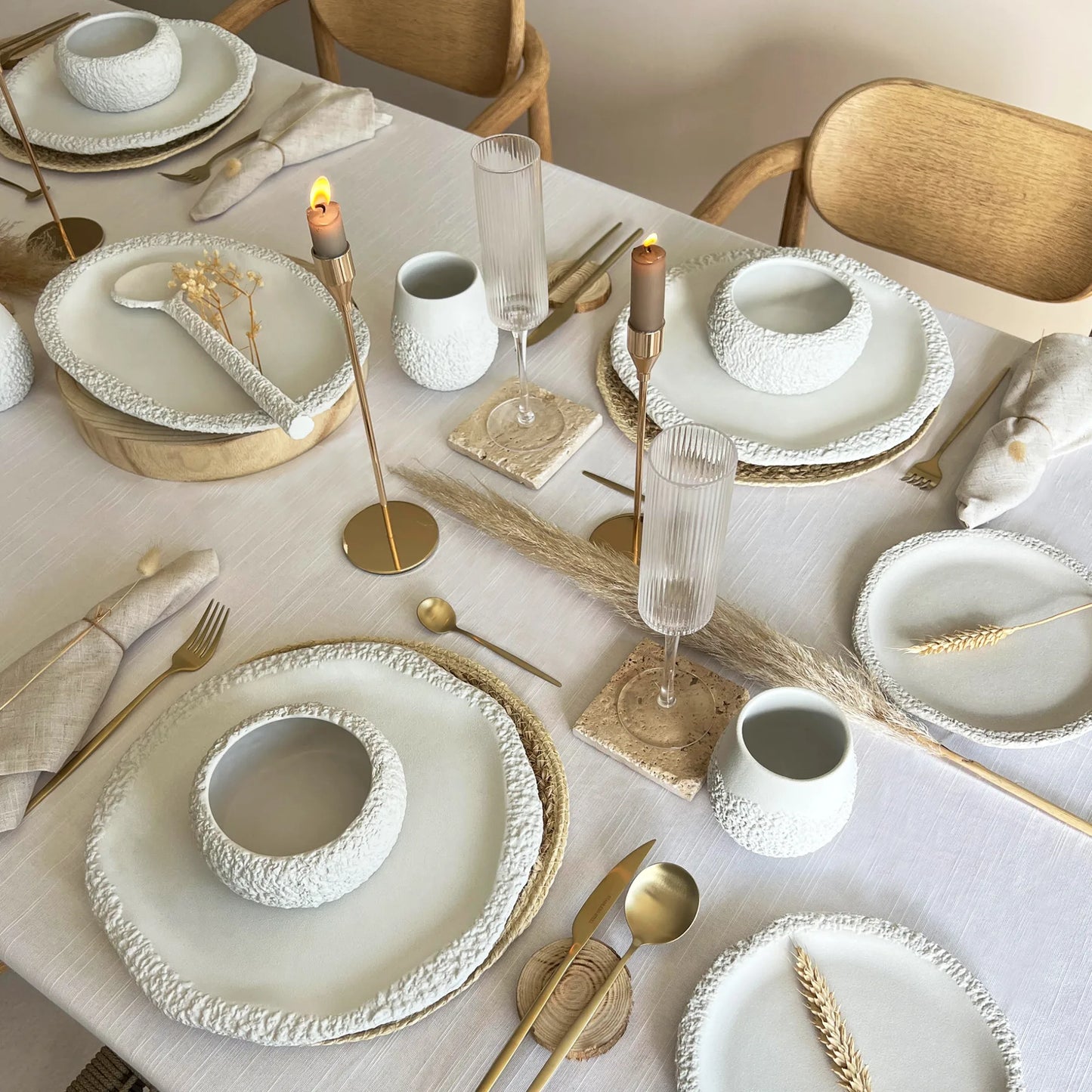 A styled dining table set with textured white ceramic dinnerware that includes plates, matching bowls, mugs,  gold-toned flatware, glass flutes, and candlesticks on a light neutral tablecloth.