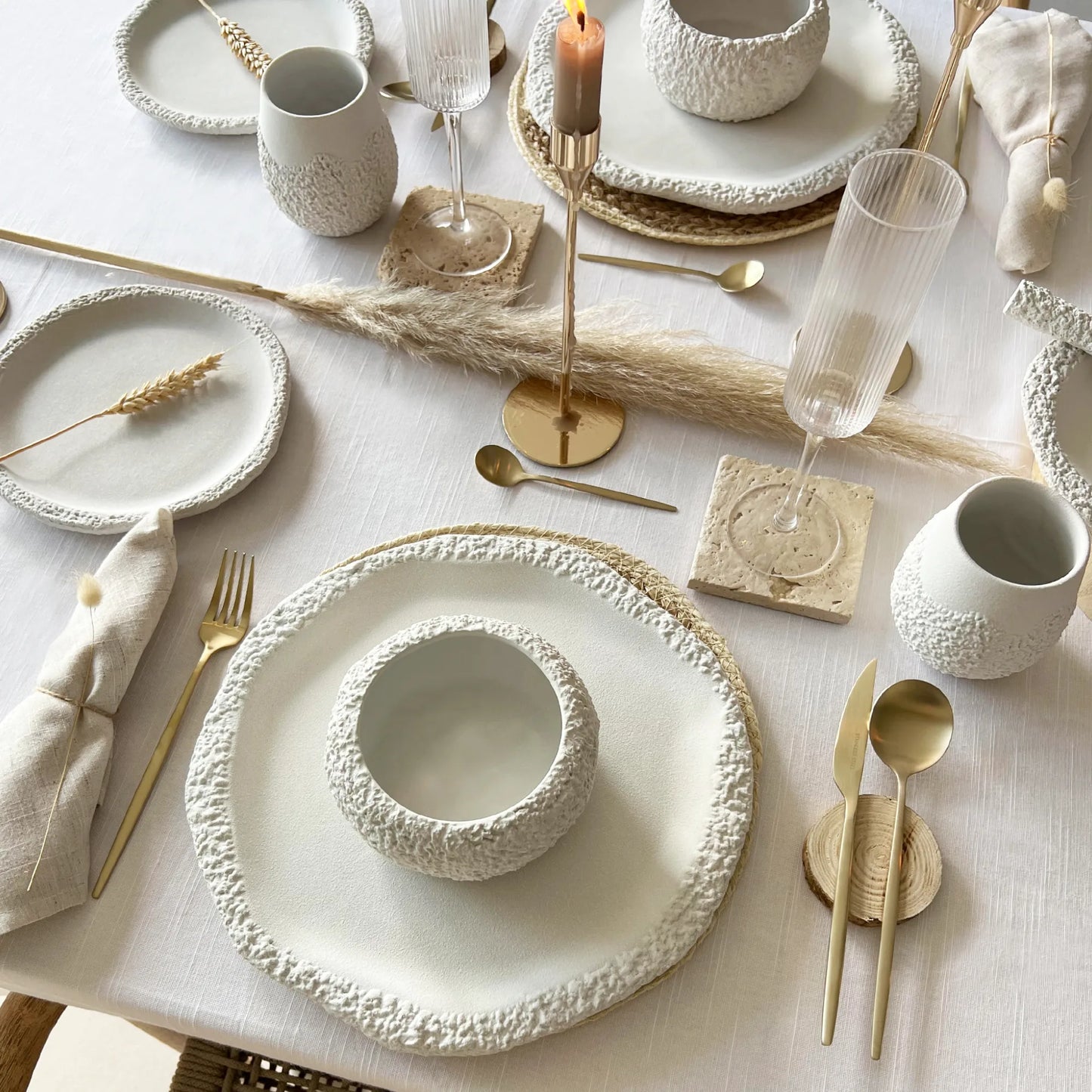 A styled table setting with textured off-white ceramic plates, bowls, cups, and glassware, mixed with gold toned flatware and candlesticks on a white tablecloth.