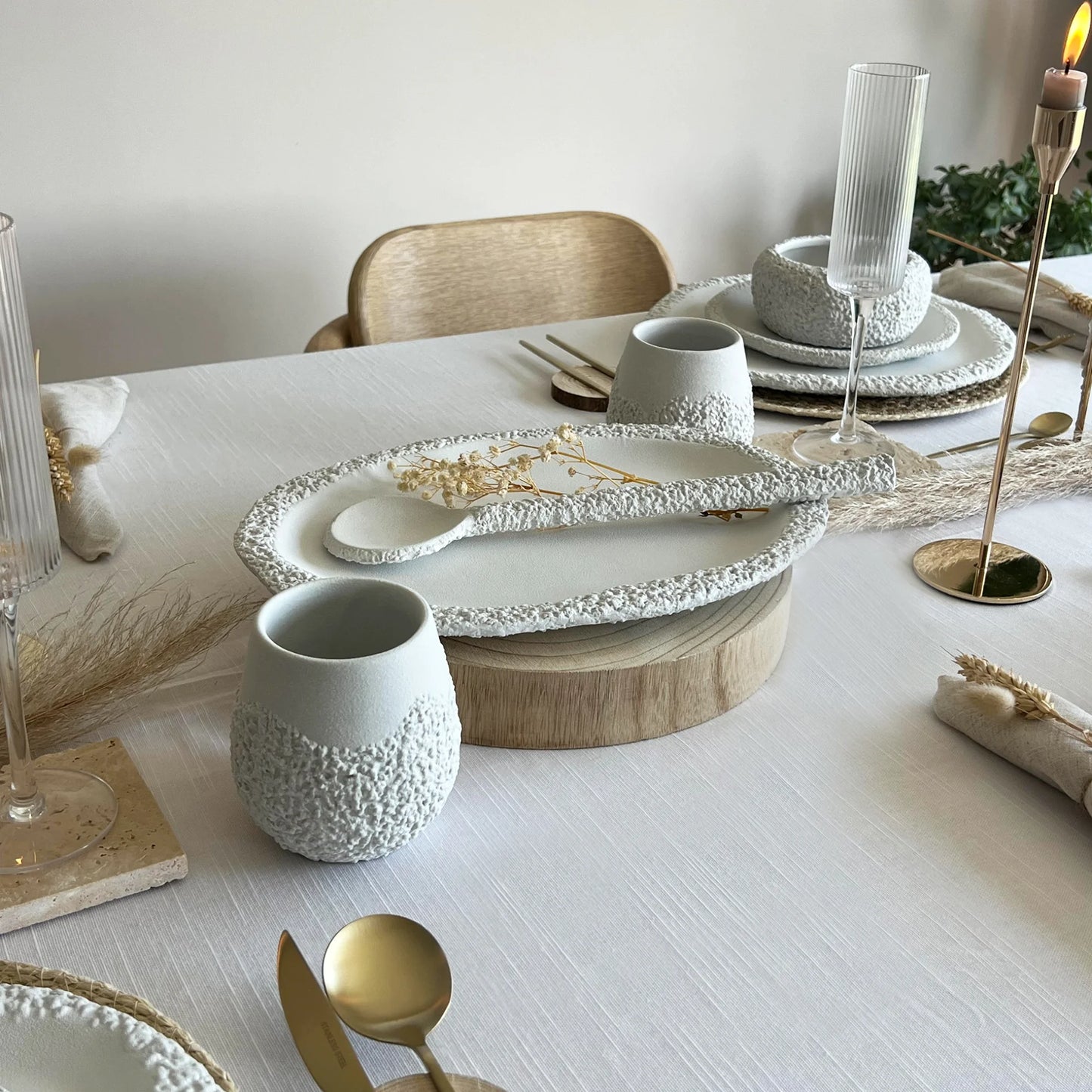 A styled dining table setting featuring textured ceramic tableware in neutral tones, including a textured oval serving platter with a matching serving spoon, a textured cup, and stacked plates, accented with gold-toned cutlery and candlesticks.