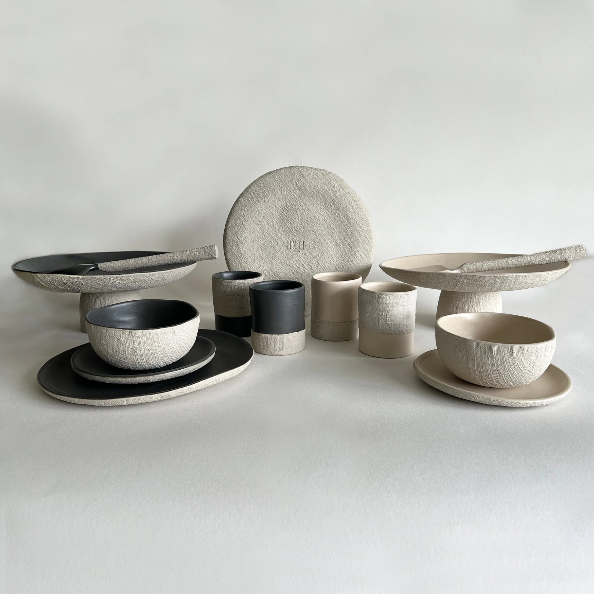 A coordinated set of handcrafted ceramic tableware in neutral tones, including bowls, cups, plates, and serving dishes in textured beige, cream, and charcoal black, arranged on a light neutral background.