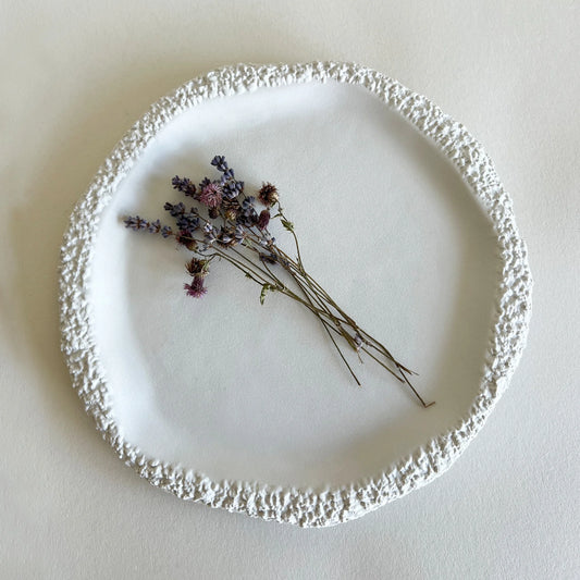 A side and dessert round white ceramic plate with a textured, raised organic rim, styled with a small dried lavender sprig on a neutral cream background.