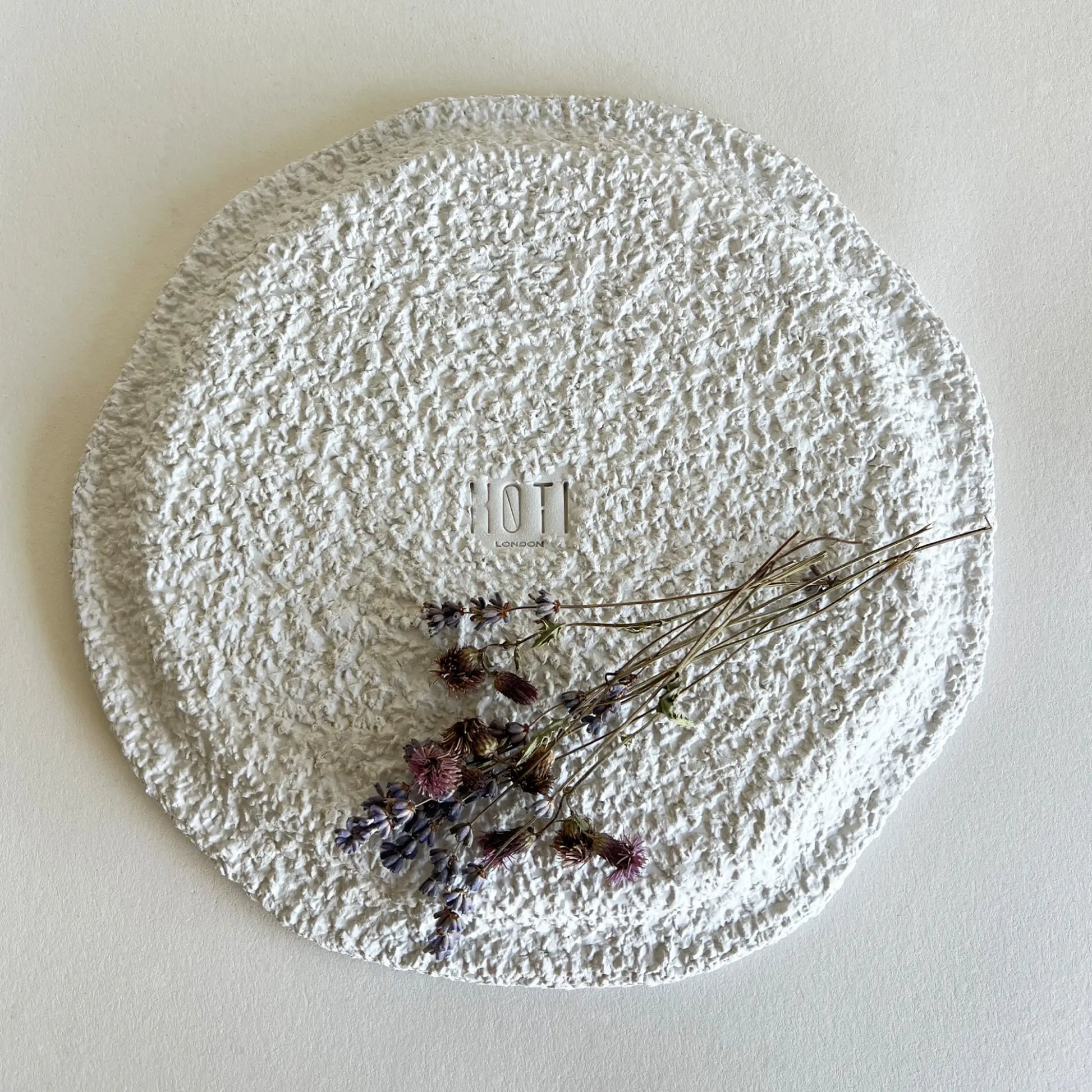 A round, large textured white ceramic plate with a rough, stone uneven finish, decorated with dried lavender sprigs.