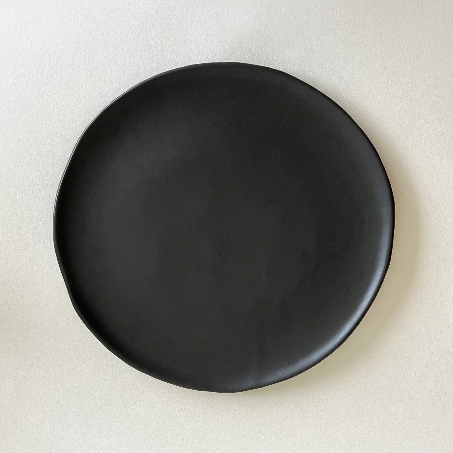 A matte black ceramic dinner plate with a slightly irregular hand-drawn edge, finished in black satin look. 