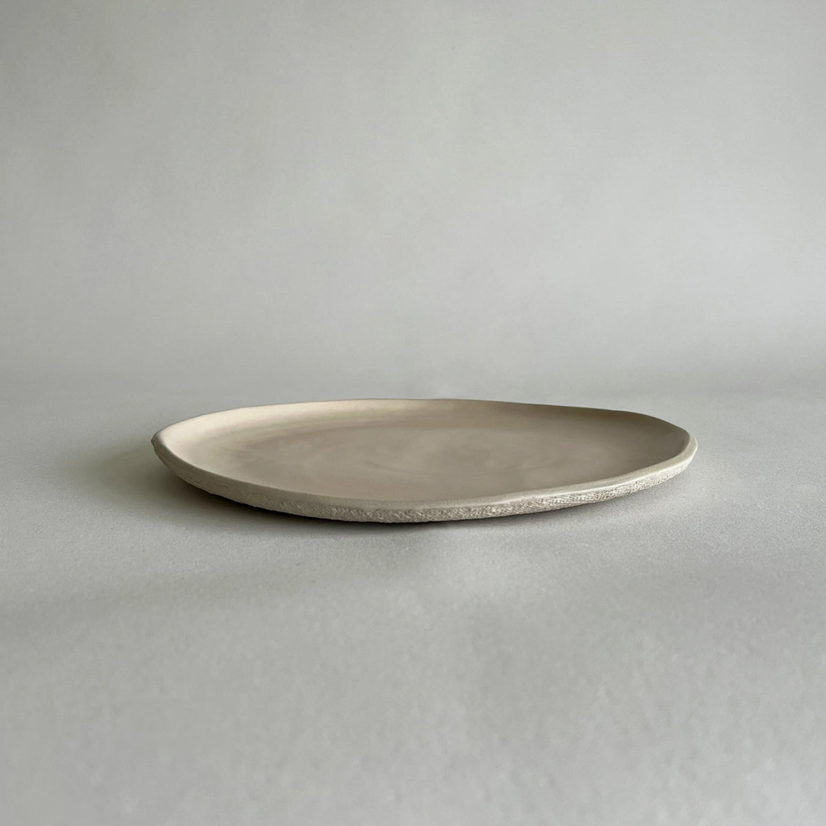 A large textured handcrafted ceramic dinner plate in a light beige or off-white glaze, photographed on a neutral light grey background.
