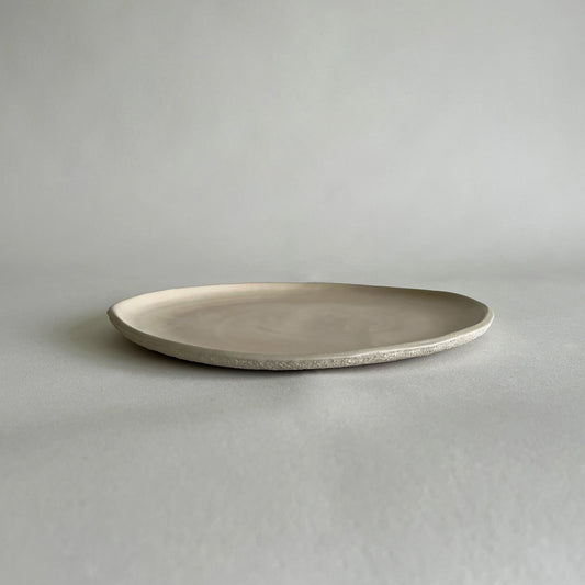 A large textured handcrafted ceramic dinner plate in a light beige or off-white glaze, photographed on a neutral light grey background.