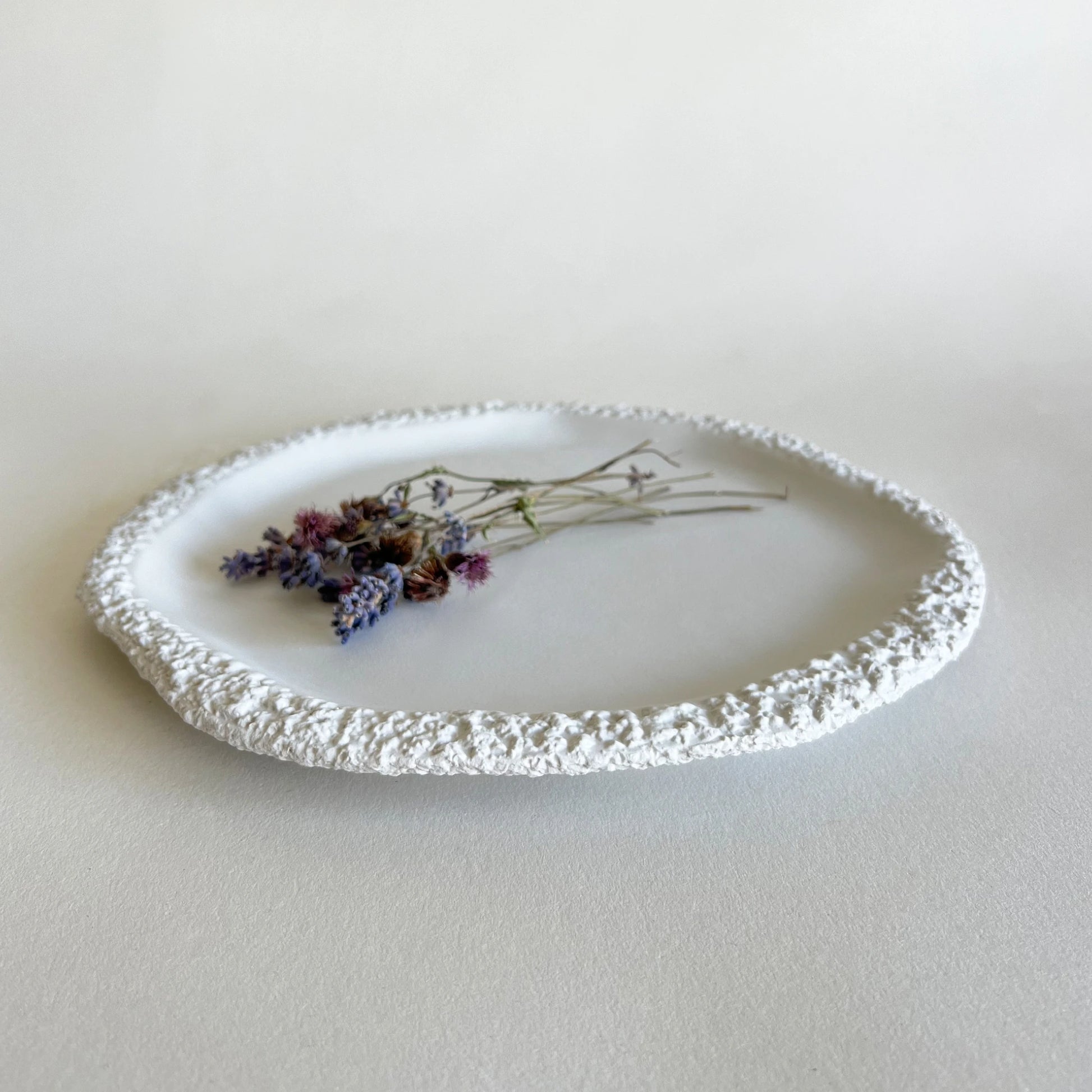 A large or main white ceramic plate with a textured, raised rim, styled with dried lavender and small flowers on a matching light beige surface.