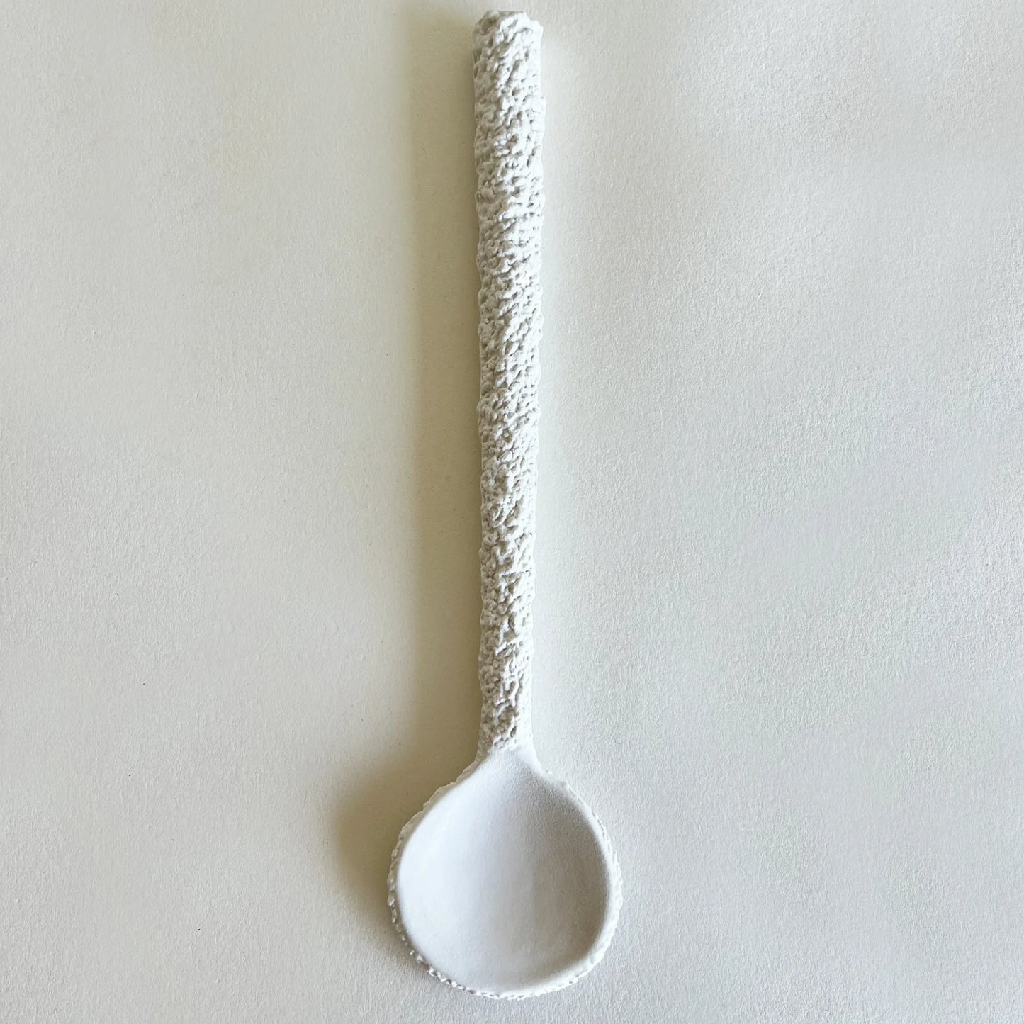 A white ceramic serving spoon with a textured, speckled handle, photographed vertically against a neutral, light beige background.