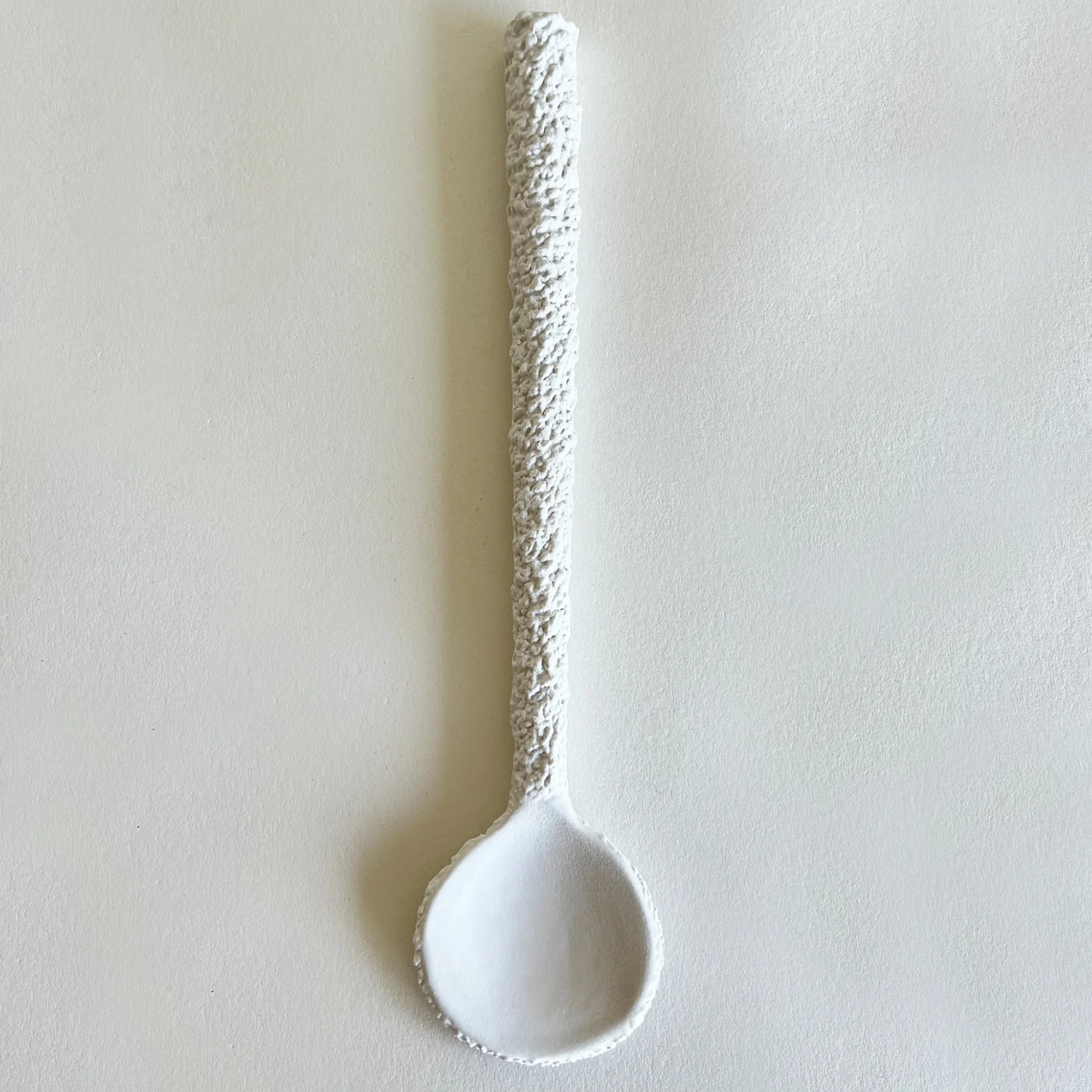 A white ceramic serving spoon with a textured, speckled handle, photographed vertically against a neutral, light beige background.