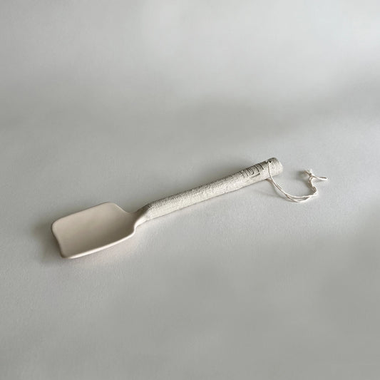 A ceramic serving spoon with a textured, speckled beige handle and a small hanging loop, photographed on a neutral light grey background.