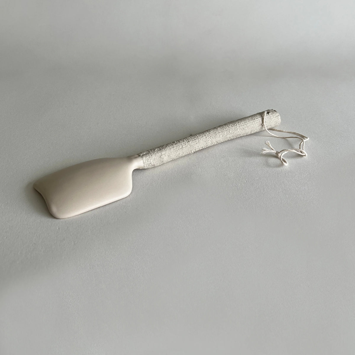 A ceramic serving spoon with a smooth, matte off-white head and a textured, organic off-white handle, finished with a matching cord loop, shown on a neutral light grey background.