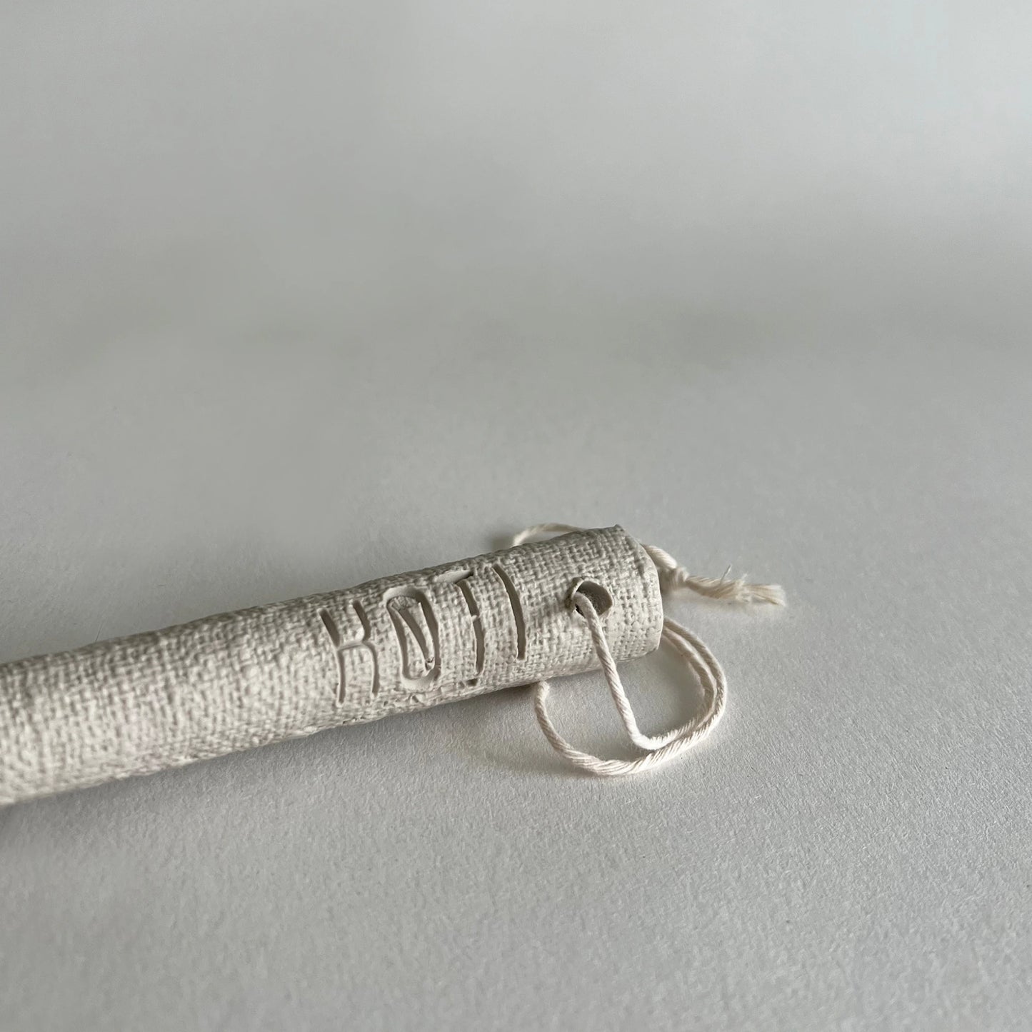 A close-up of a cylindrical textured beige serving spoon handle, attached to a small loop of white cord, photographed on a light grey background.
