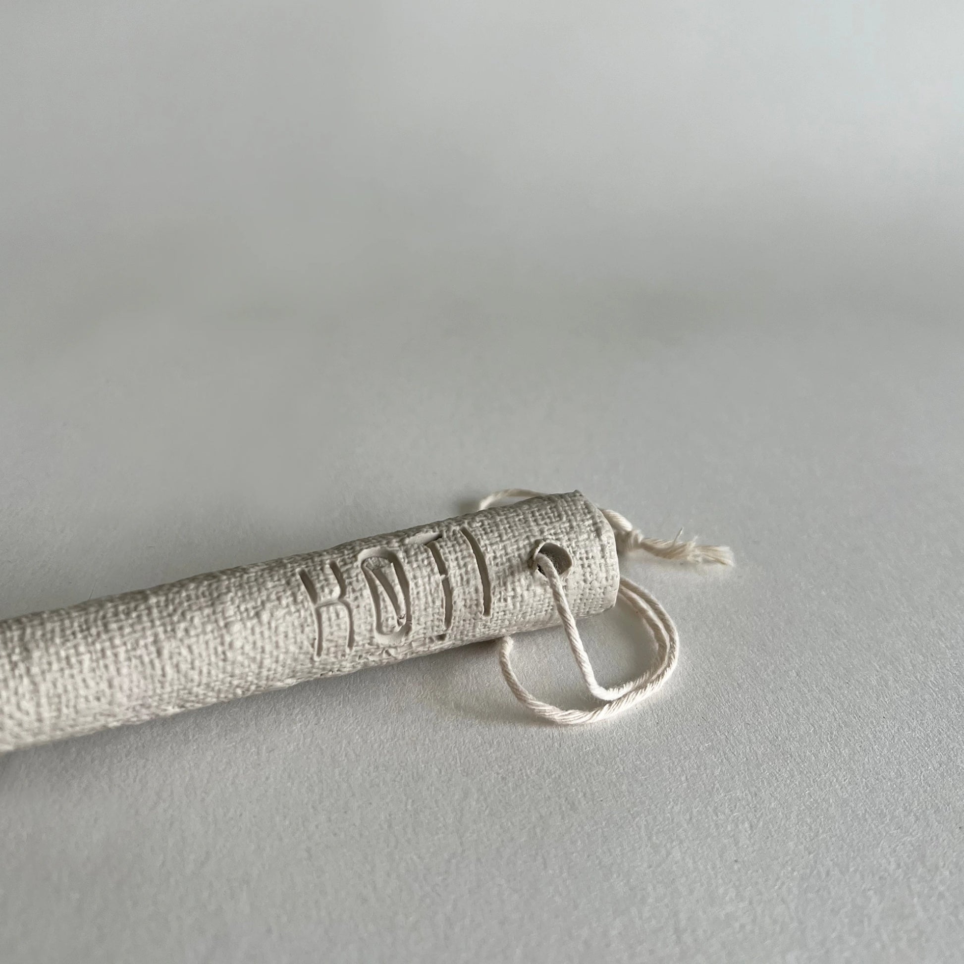 A close-up of a cylindrical textured beige serving spoon handle, attached to a small loop of white cord, photographed on a light grey background.