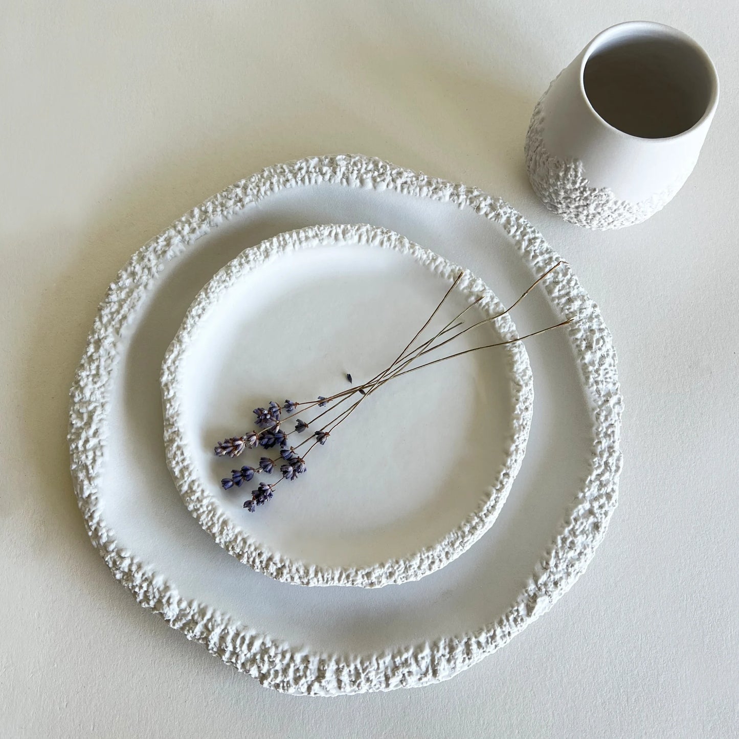 two textured white ceramic plates in nested round sizes with a small matching white cup, styled with dried lavender on a light beige surface.