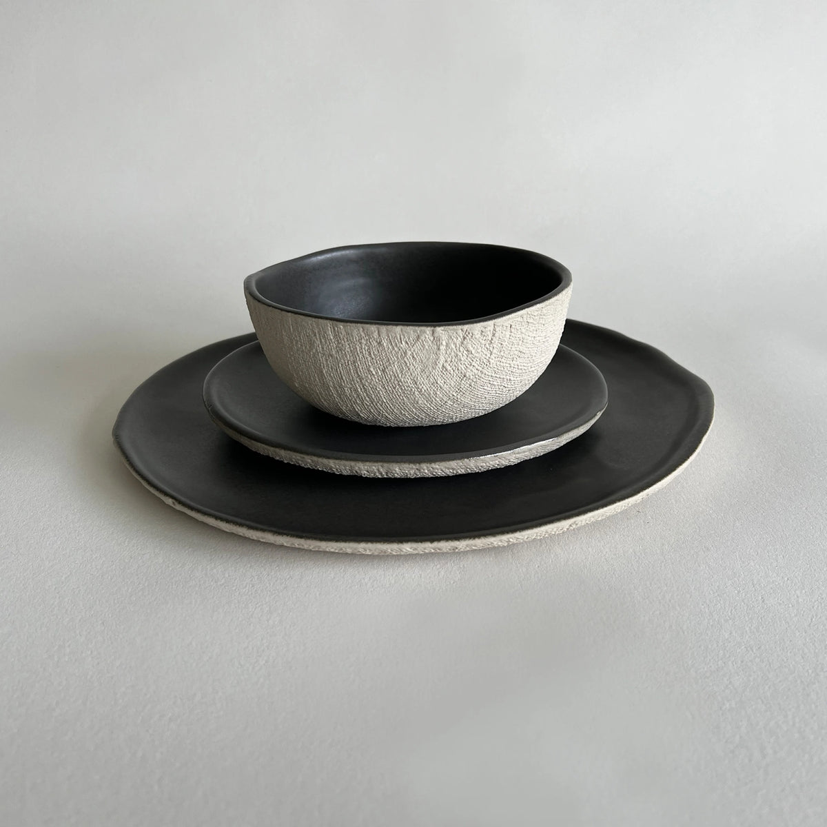 A ceramic tableware set featuring a textured dark grey bowl, matching grey side plate, and a dinner plate with a contrasting textured beige rim.