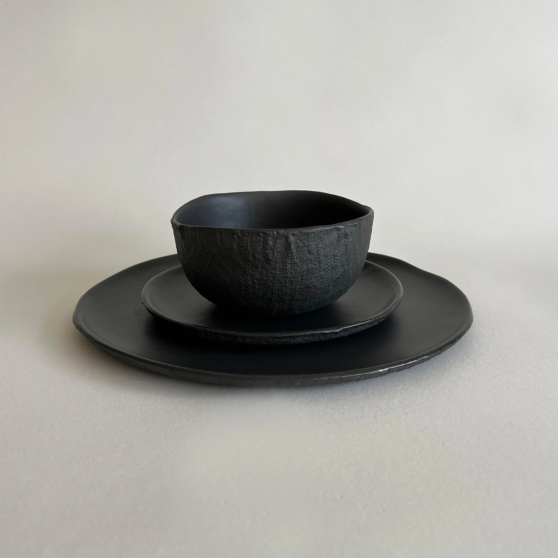 A black textured ceramic bowl set with large dinner plate and side plate, showcasing organic linen texture.