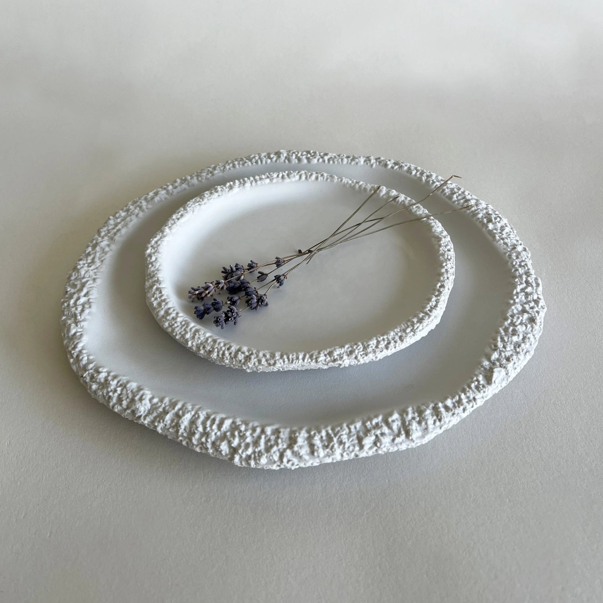 Two nested round ceramic plates with textured, organic raised edges in white, styled with a small sprig of dried lavender on a neutral light grey background.