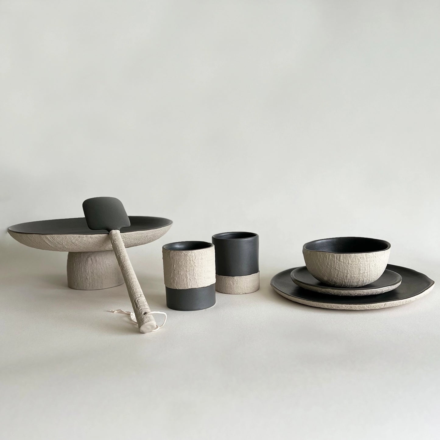 A coordinated ceramic tableware set in matte charcoal and textured warm beige, including a pedestal platter with a serving utensil, two coffee cups, matching bowls, dinner plate and salad plate.