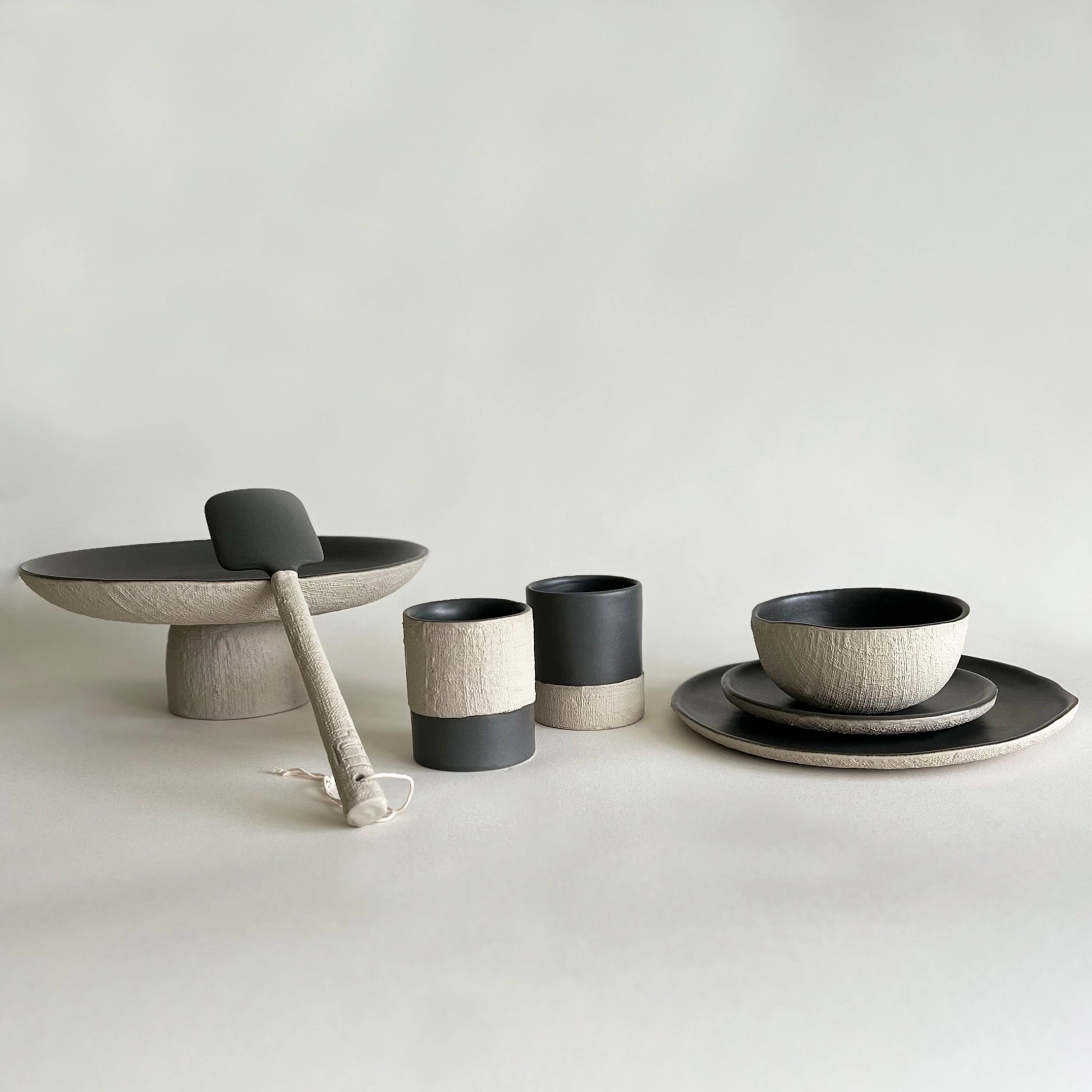 A coordinated ceramic tableware set in matte charcoal and textured warm beige, including a pedestal platter with a serving utensil, two coffee cups, matching bowls, dinner plate and salad plate.