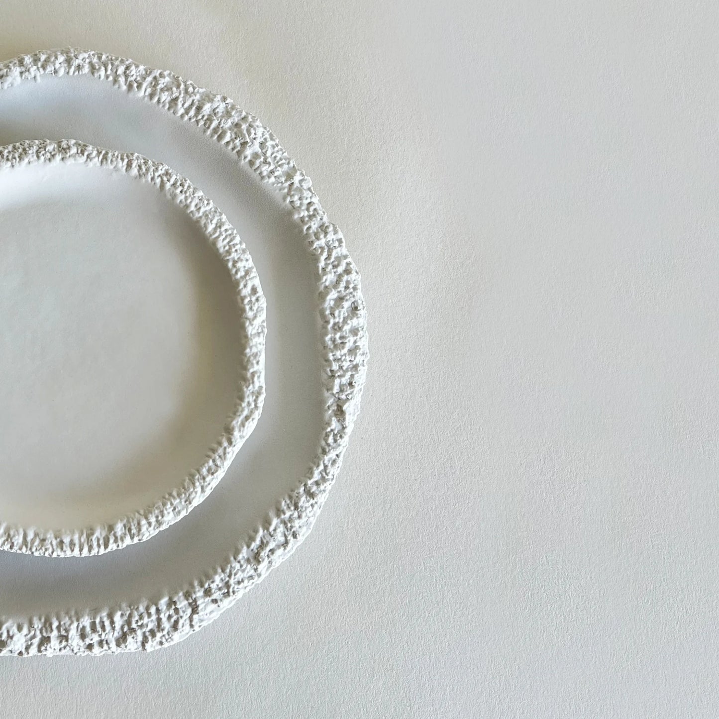 Two textured white ceramic plates with raised, raw edges, arranged concentrically on a light grey surface.