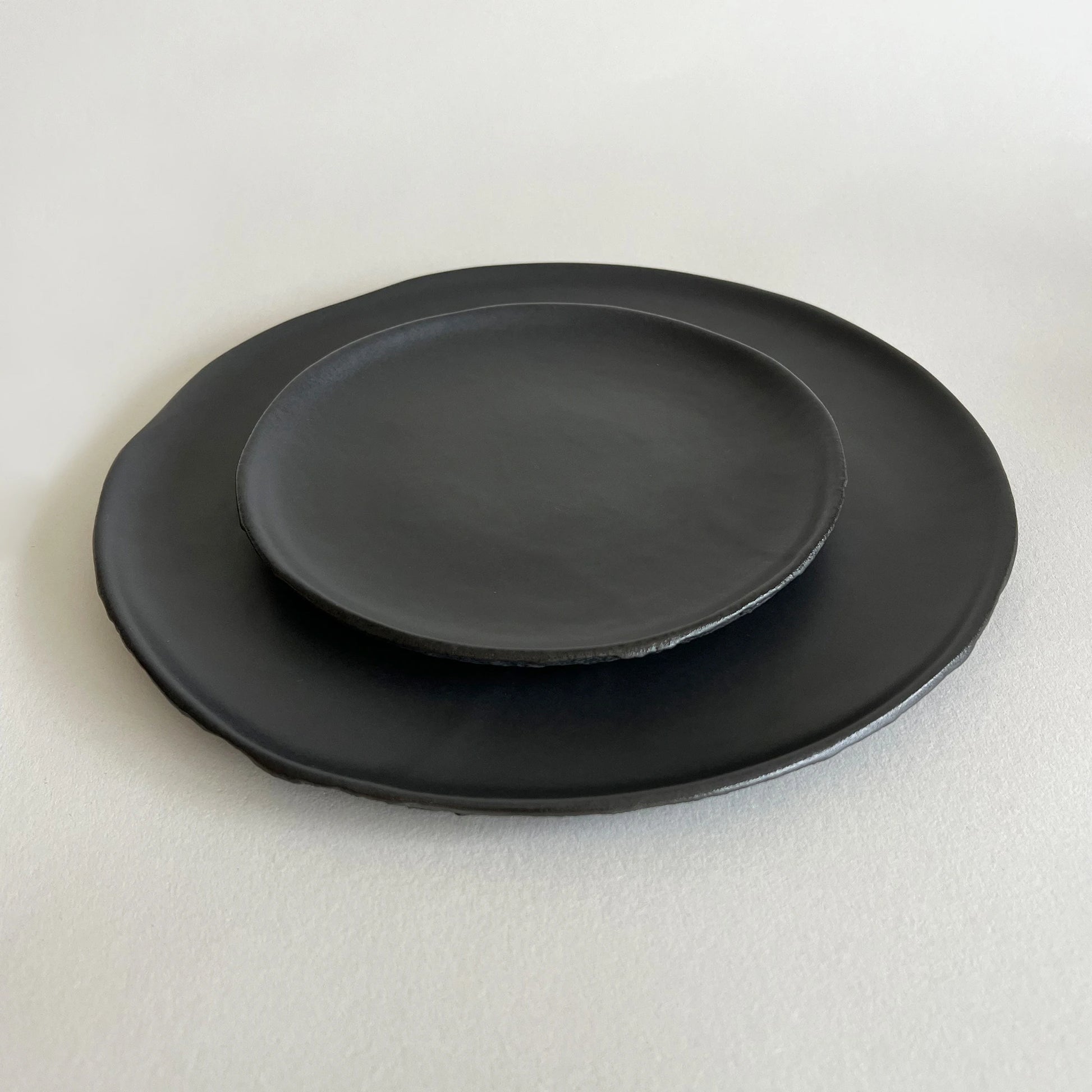 A set of two handmade ceramic plates, includes one large dinner plate in matte black and one salad plate in matte black, both finished with raw texture.