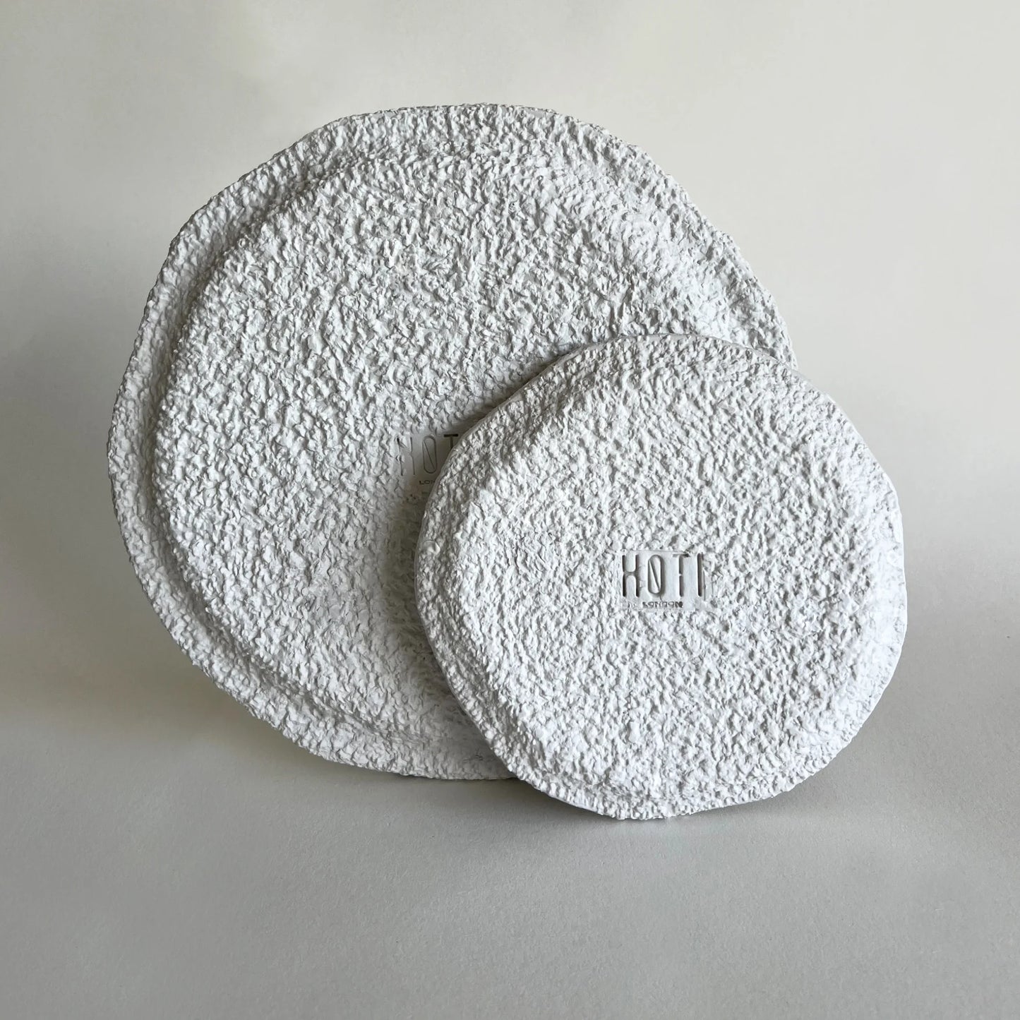 Two round, textured white plates with a rough, stucco-like finish, one larger plate standing upright behind a smaller round plate lying flat. 