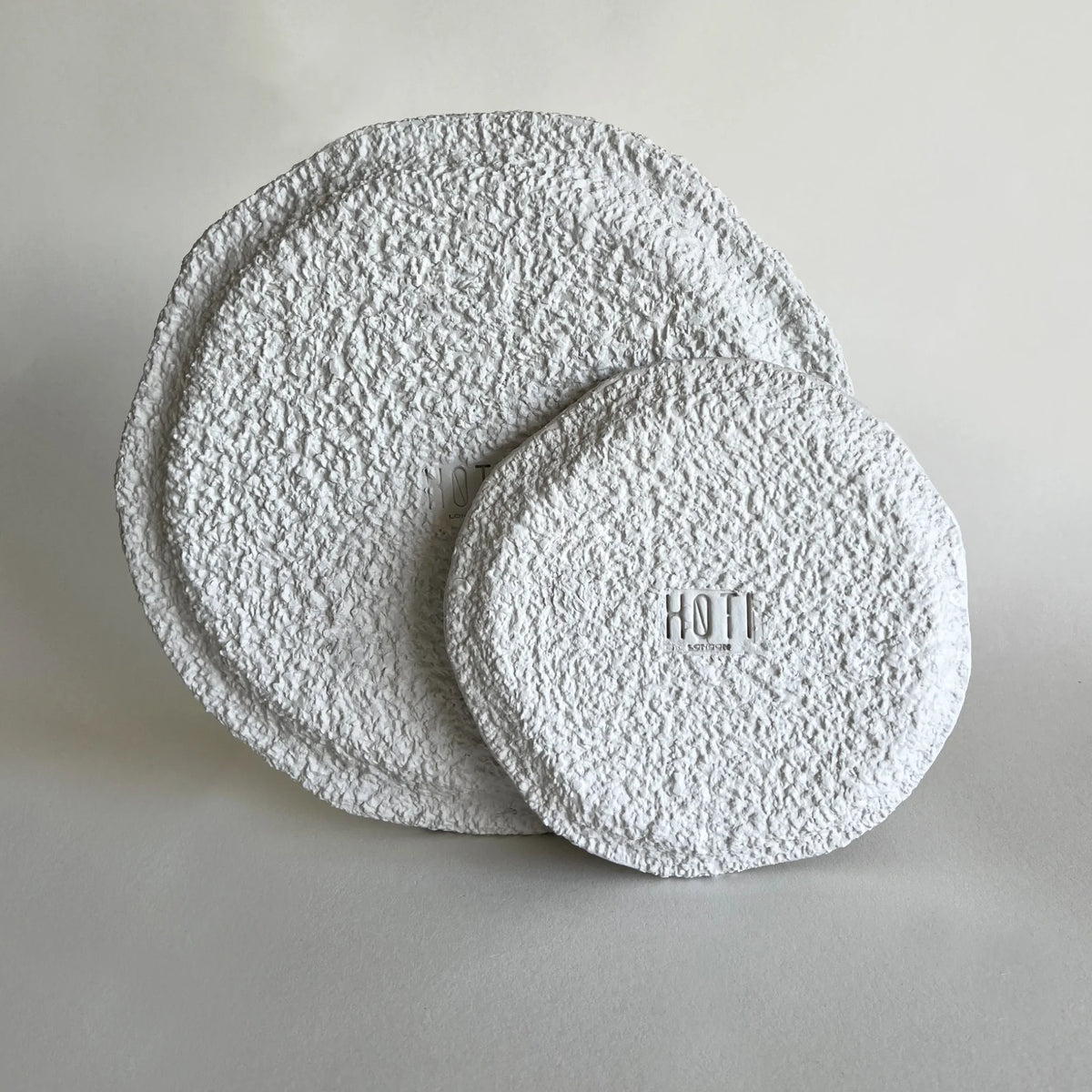 Two round, textured white plates with a rough, stucco-like finish, one larger plate standing upright behind a smaller round plate lying flat. 