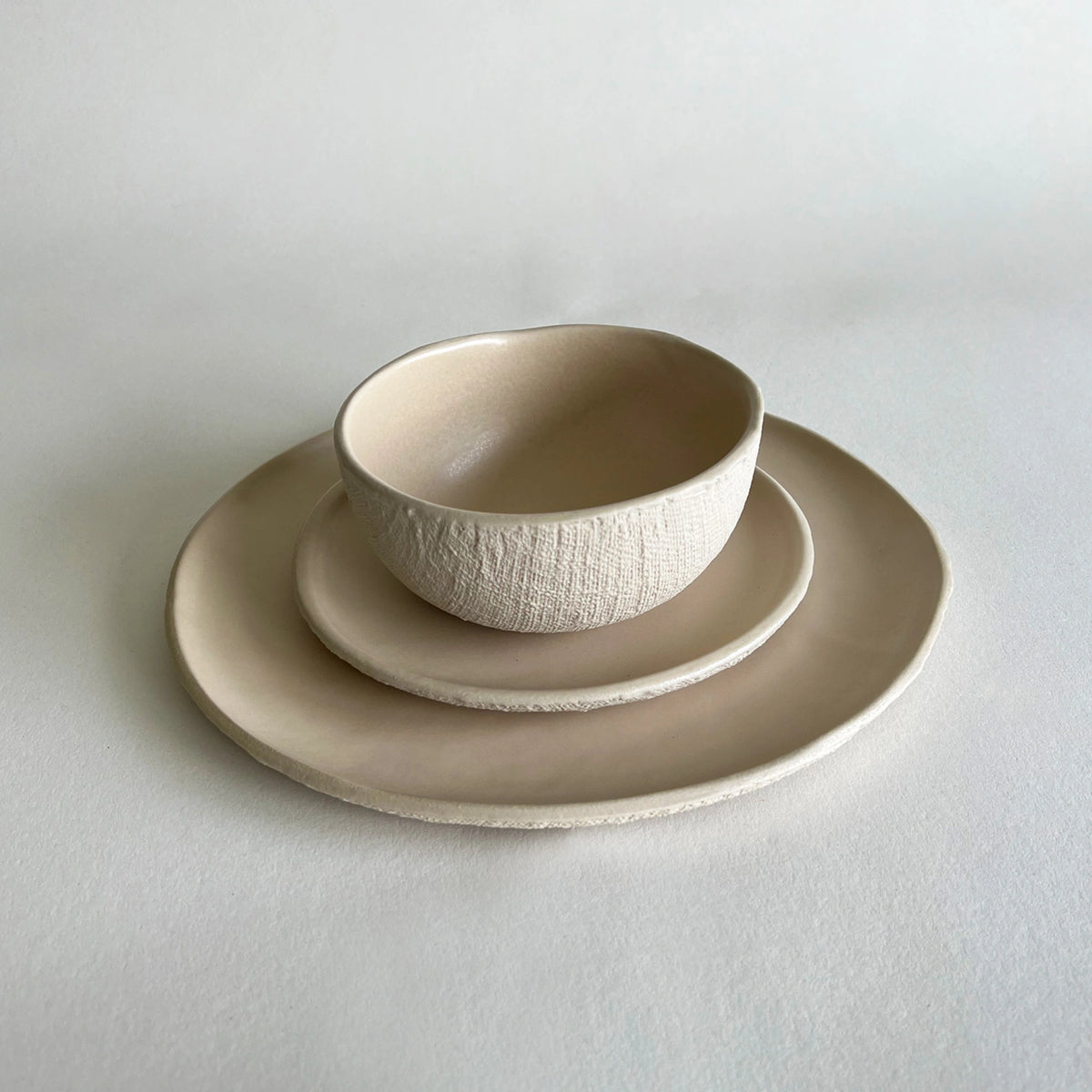 A ceramic bowl with a textured exterior and smooth glazed interior, nested on a matching side plate, which sits on a large round platter, all in a warm off-white/cream tone against a neutral light grey background.