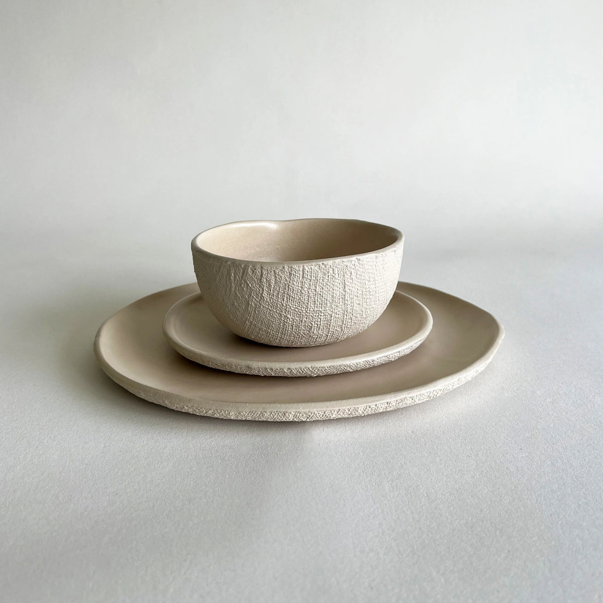 A minimalist ceramic tableware set  in beige colour featuring a soup or ramen textured bowl, a matching dessert plate, and a large dinner plate in a warm cream-beige colour, arranged on a neutral light grey background.