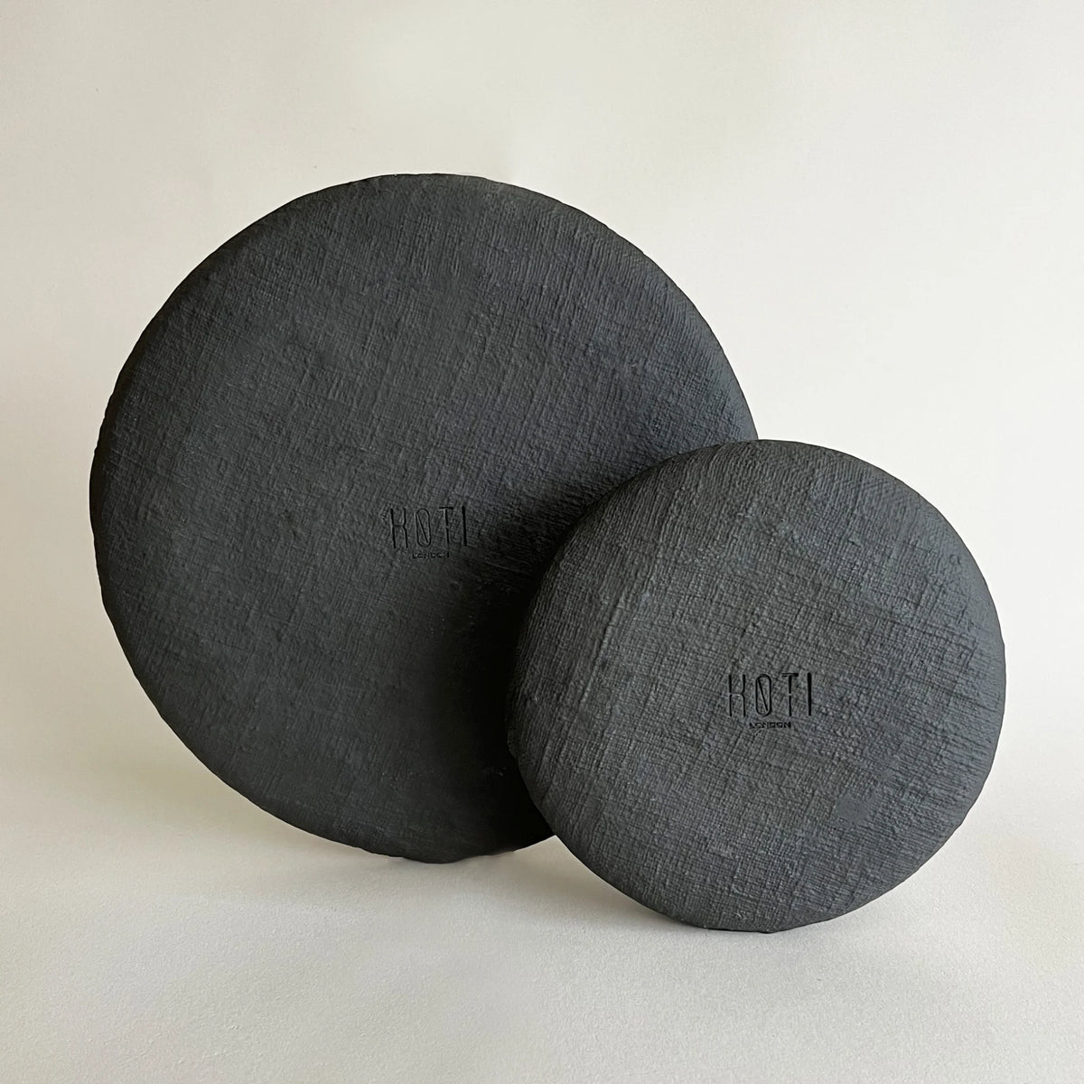 Two round matte black plates, one is large dinner plate followed by dessert plate, showcasing uneven raw textured finish.  