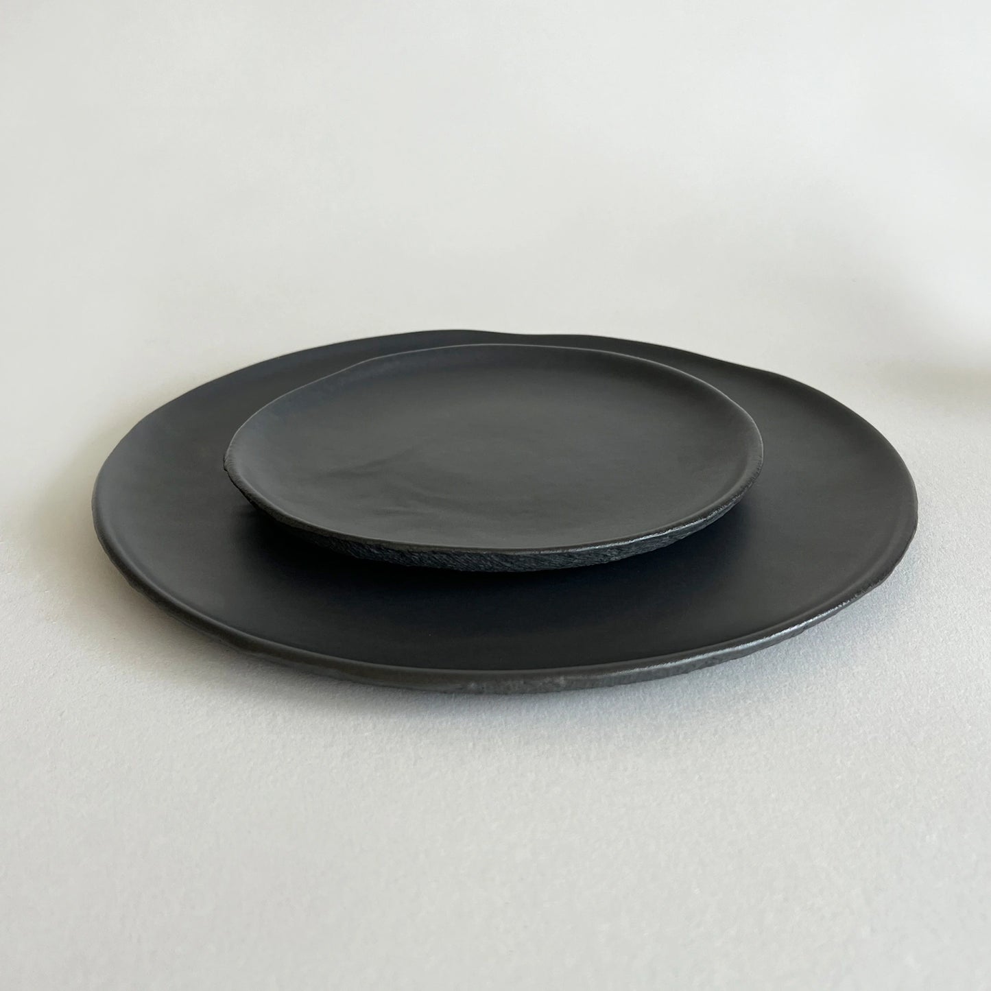 Set of two matte black ceramic plates, one is large pizza plate and the other is matching side plate, both with organic raw texture.