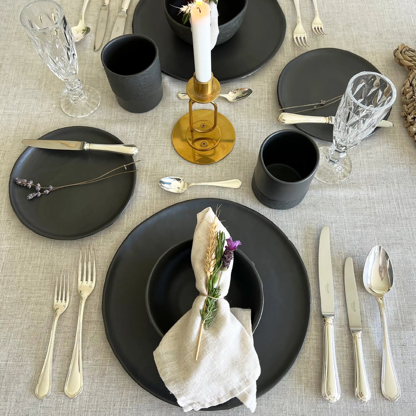 A styled table setting with matte black dining plates, matching black cups, silver flatware, clear cut-glass goblets, a gold candle holder with a white taper, and a beige linen napkin with a small purple flower, all arranged on a light beige textured tablecloth.